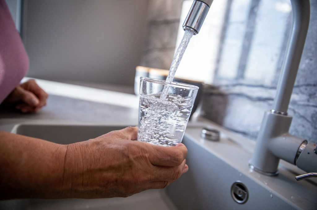 A person holding a clear glass under a kitchen faucet as water flows in, capturing the moment the glass is being filled with tap water.