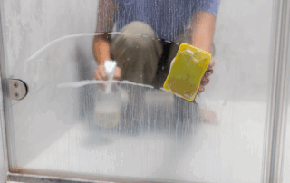 A person cleaning a foggy glass shower door from hard water residue with a yellow sponge and a spray bottle. The image shows soap and water streaks being wiped away, revealing a clearer section of the glass.