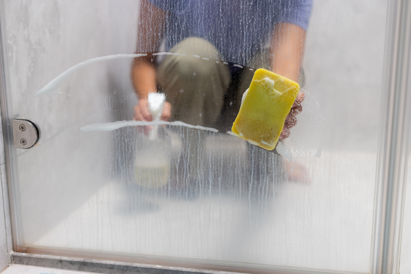 A person cleaning a foggy glass shower door from hard water residue with a yellow sponge and a spray bottle. The image shows soap and water streaks being wiped away, revealing a clearer section of the glass.