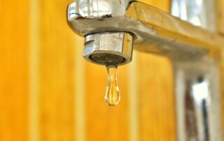 Close-up of a chrome faucet with a single droplet of water hanging from the spout against a blurred yellow background.