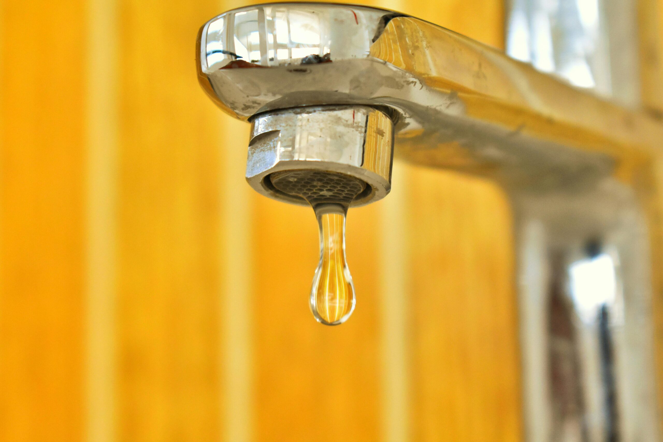 Close-up of a chrome faucet with a single droplet of water hanging from the spout against a blurred yellow background.